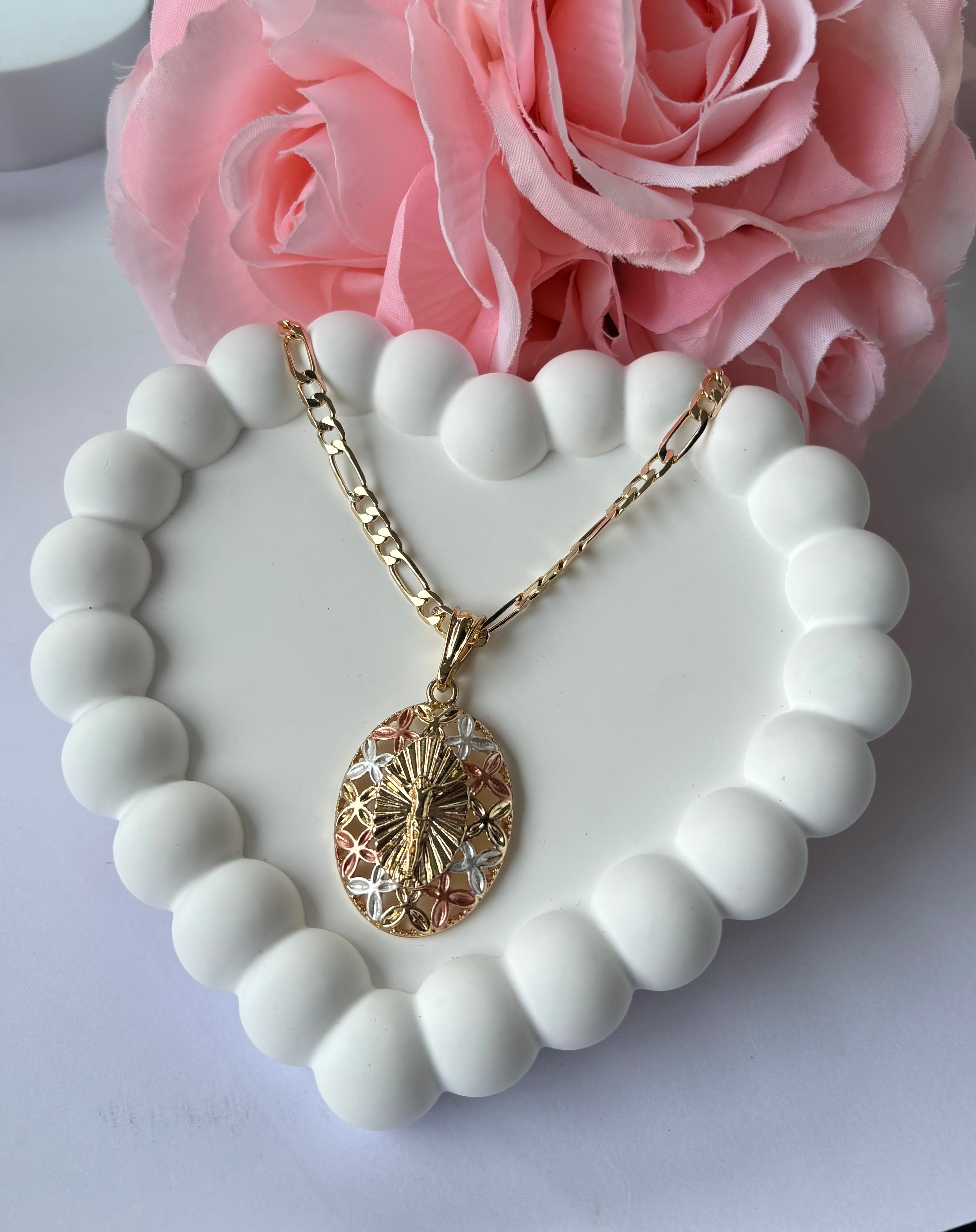 Tri-Color Cross Figaro Necklace with oval pendant featuring gold, silver, and rose-toned details, displayed on a white beaded heart tray with pink roses.
