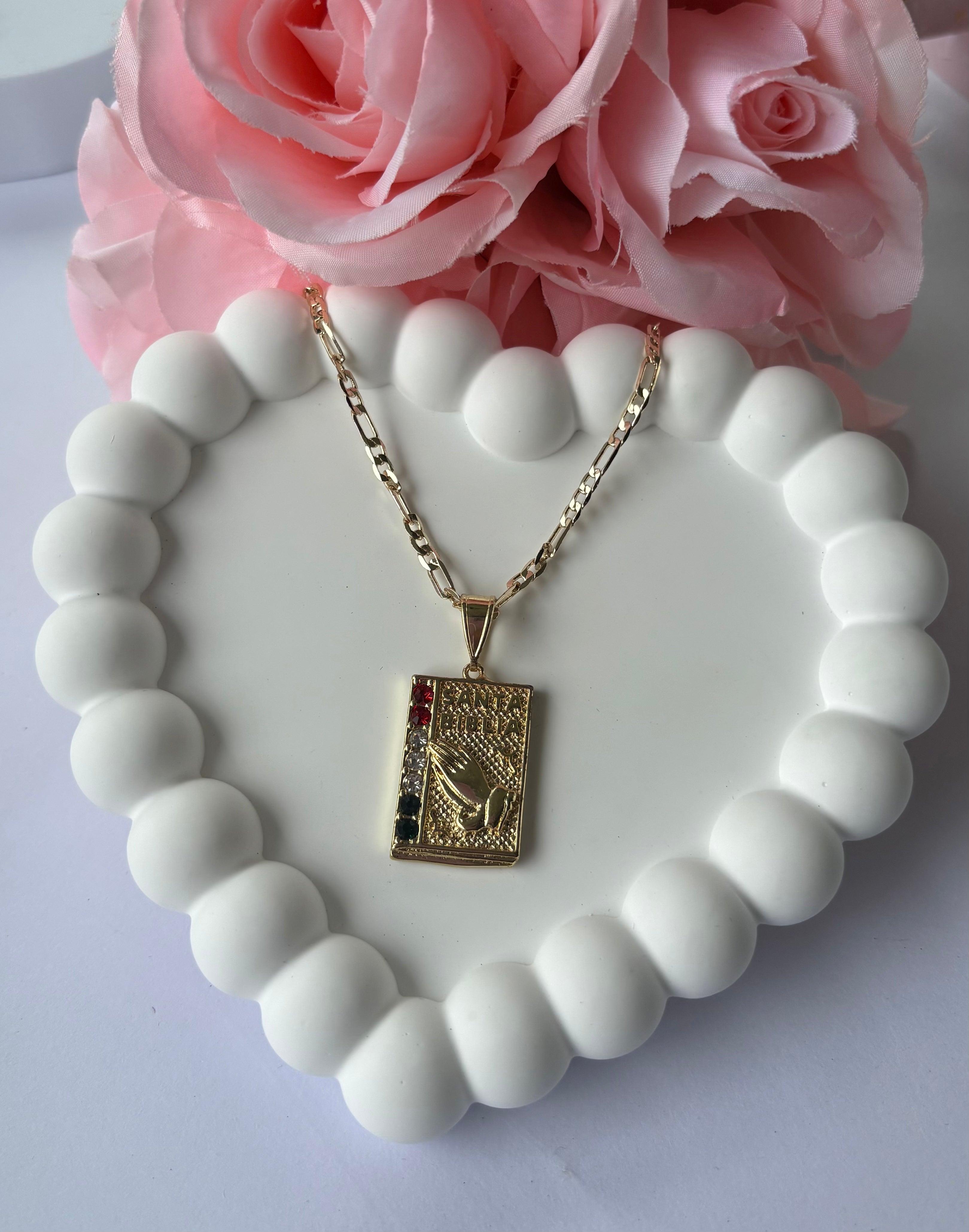 Gold plated Bible Necklace with praying hands design and subtle red, white, and blue accents, displayed on a white beaded heart tray with pink roses.