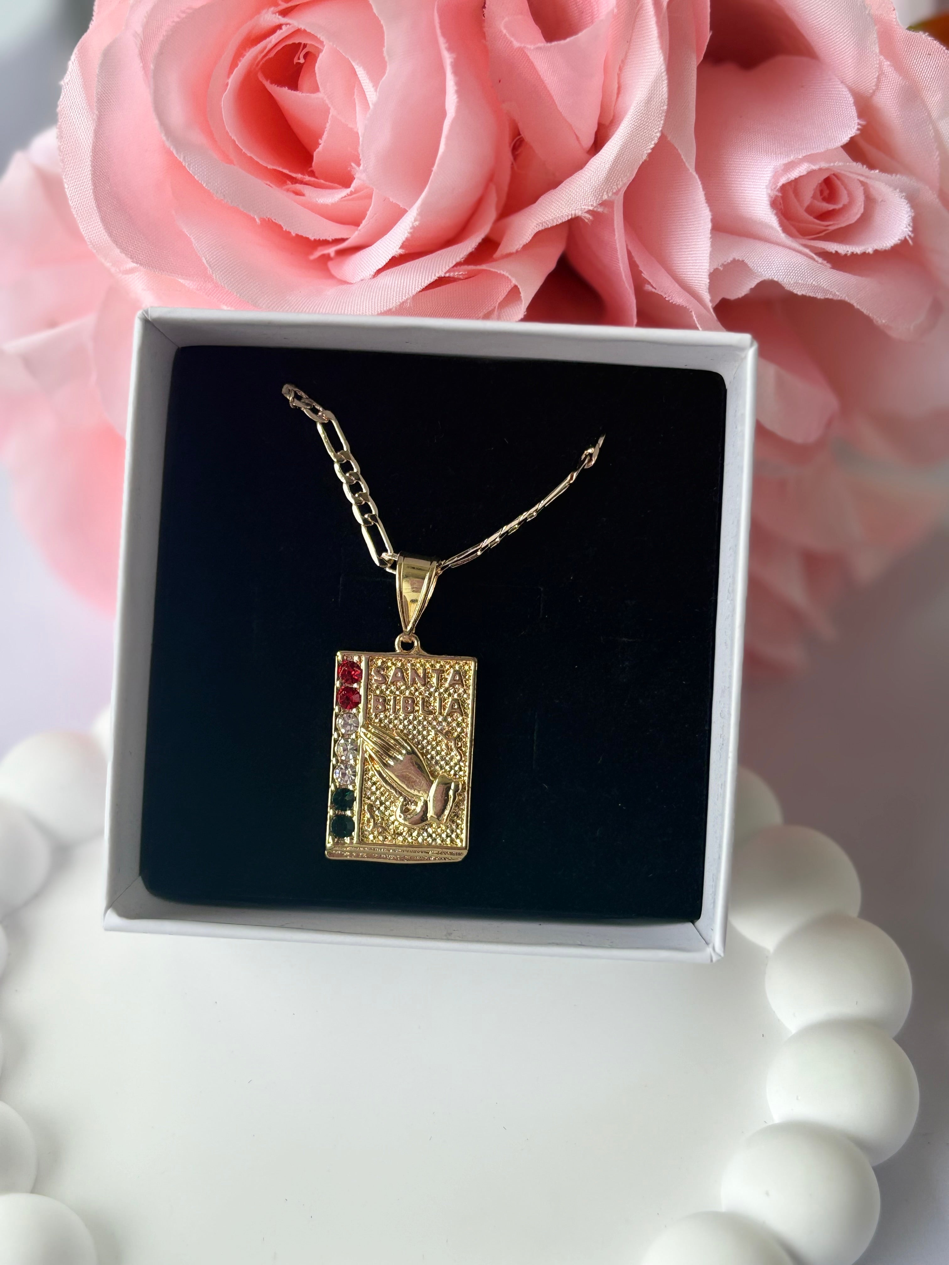 Gold plated Bible Necklace with praying hands design and subtle red, white, and blue accents, displayed on a white beaded heart tray with pink roses.