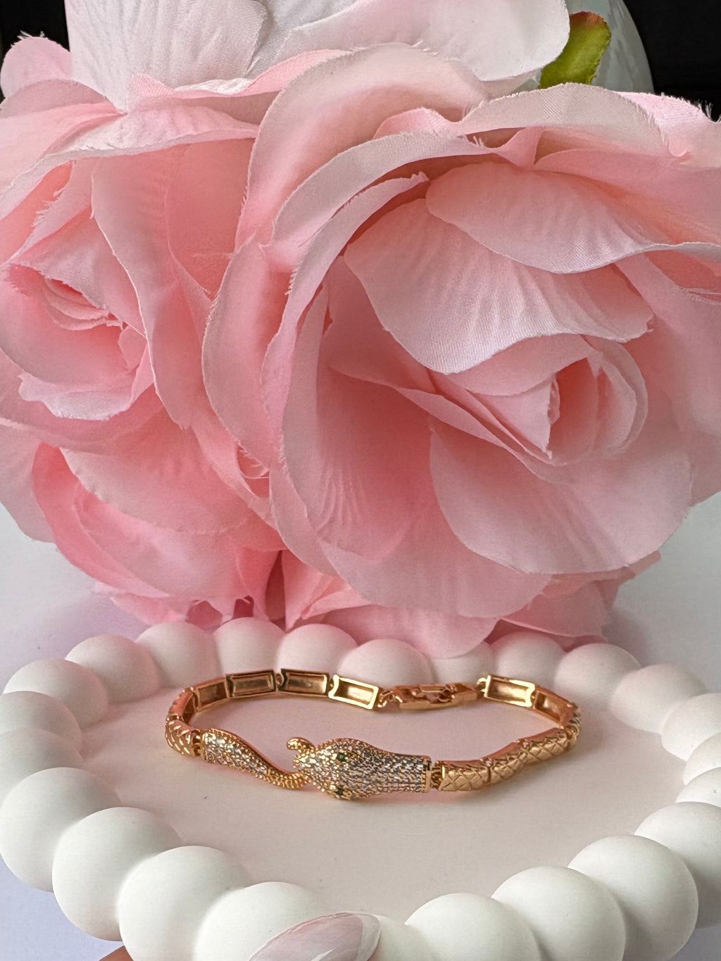 Hand holding a white beaded bracelet with gold accents next to pink flowers on a light background
