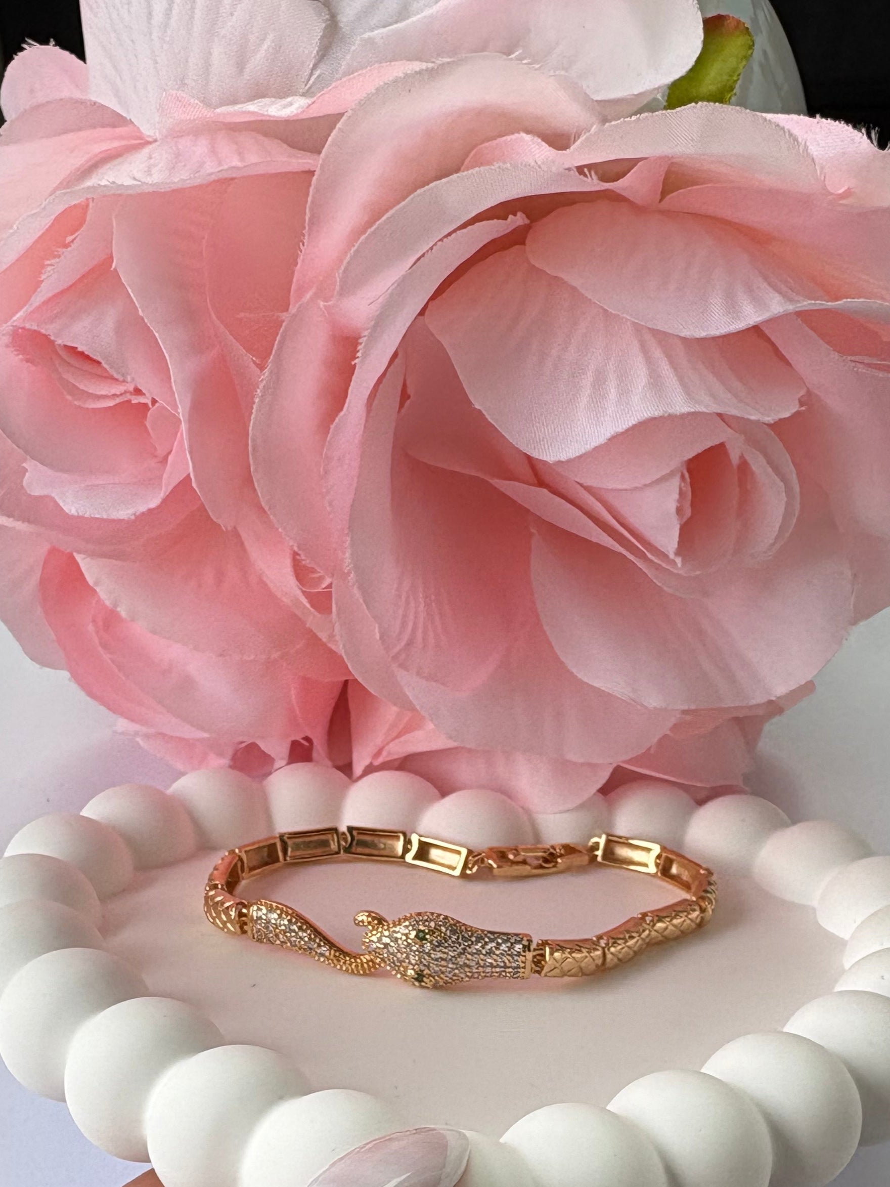 Hand holding a white beaded bracelet with gold accents next to pink flowers on a light background