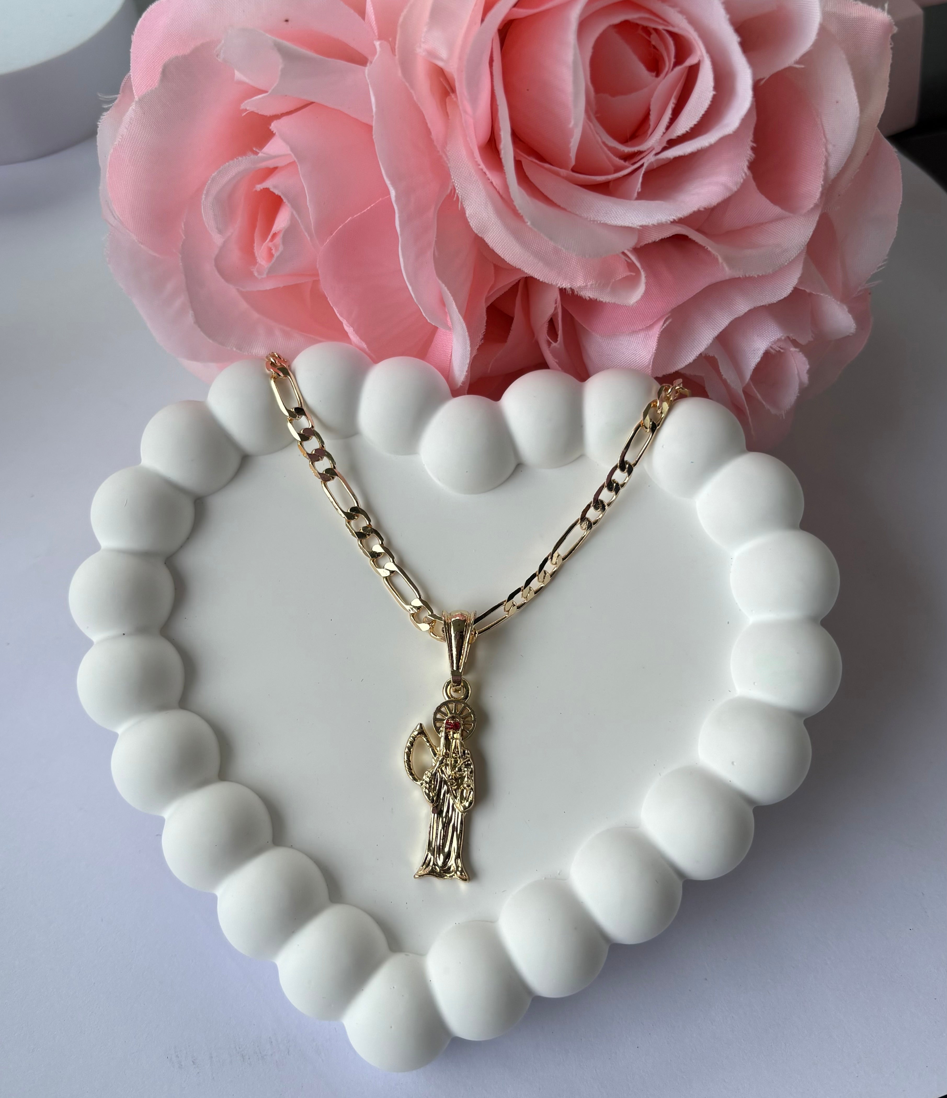 Gold plated Santa Muerte necklace with detailed pendant, displayed on a white beaded heart tray with pink roses.