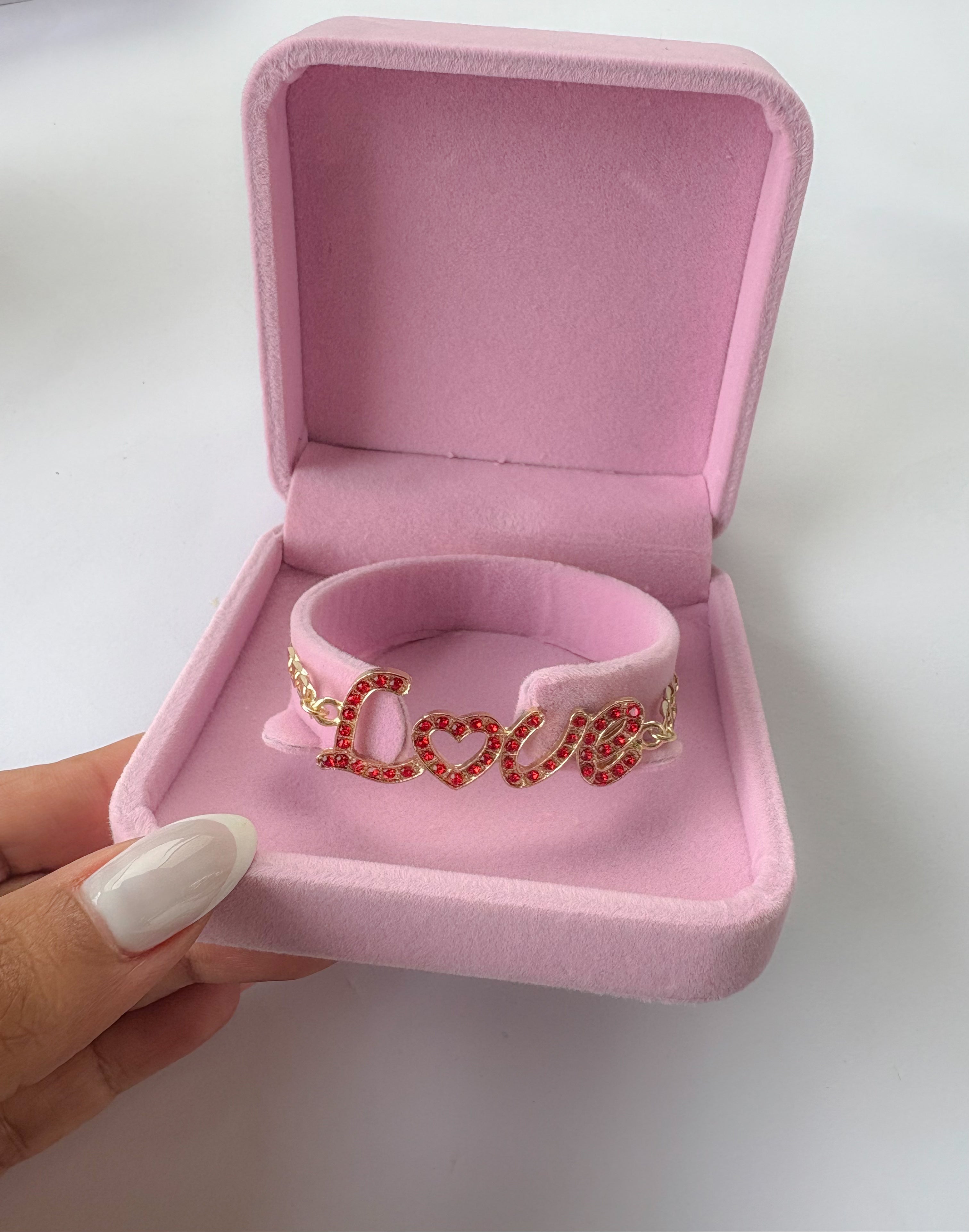 Gold plated Love Bracelet with sparkling red stones, worn on wrist with white manicure, displayed with pink roses in background.